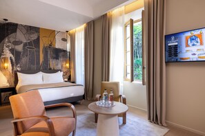 A hotel room with a bed, a chair, a table with bottles, a painting on the wall, and a TV displaying a welcome message.