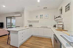 Een moderne keuken met witte kasten, een houten aanrechtblad en een inbouwvaatwasser. Drie ingelijste foto’s hangen aan de muur, en een raam zorgt voor natuurlijk licht.