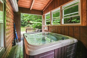 Une cabane en bois dotée d’un jacuzzi à moitié vitré, d’une terrasse avec mobilier d’extérieur et offrant une vue sur une zone boisée.