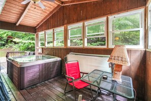 A wooden deck with a hot tub, a table with a chair, and a window with a view of greenery. - Hocking Hills Majestic Retreats - Nancy's Cabin (Rockbridge)
