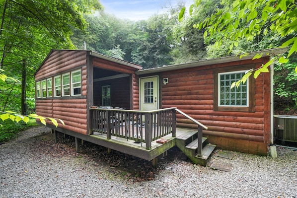 A log cabin with a porch and a small deck, surrounded by dense forest.