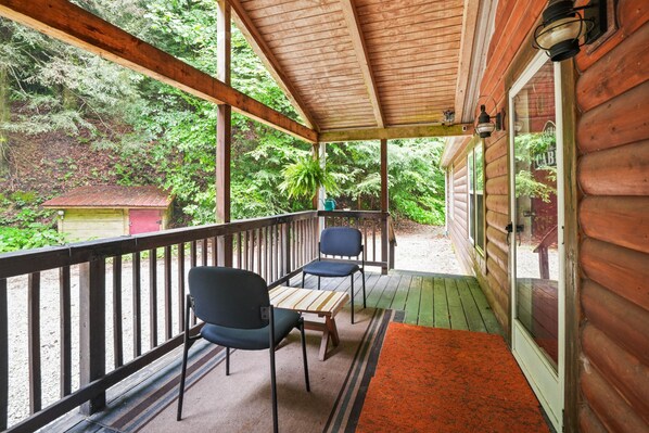 A wooden porch with two chairs and a table, overlooking a forested area.