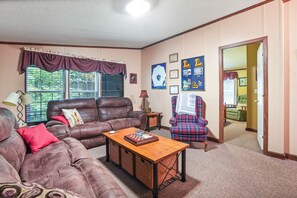 Ein Wohnzimmer mit einer braunen Ledersofa, einem Holztisch und einem karierten Sessel. An der Wand hängen Bilderrahmen und ein Fenster mit Jalousien.