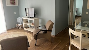 Una sala de estar con televisor sobre un mueble, una silla, un ventilador pequeño y una mesa de vidrio.
