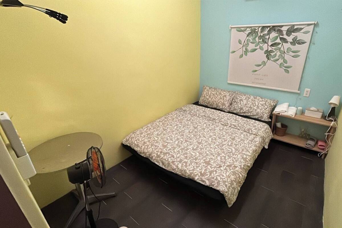 A small, well-lit room with a bed, a heater, a shelf with toiletries, and a wall-mounted plant.