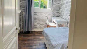 Un dormitorio con pared de ladrillo, una ventana con vista a follaje, una cama blanca, una mesita y una silla.