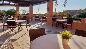 Un patio exterior con mesas y sillas redondas, plantas en macetas y vistas de la ciudad al fondo.