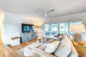 Una sala de estar moderna con una pared de ladrillo blanco, un televisor de pantalla plana, un mueble de madera, un sofá, una mesa de centro y un ventanal con vistas a muebles de exterior.