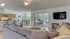 A spacious living area with a gray sofa, a ceiling fan, a kitchen with wooden cabinets, and a large TV on the wall. - Pensacola Home w/ Large Yard: 4 Mi to Beach (Pensacola)
