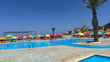 A swimming pool with multiple colorful umbrellas, lounge chairs, and a palm tree.