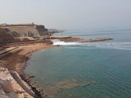 Zona costera con costa rocosa, agua cristalina y un edificio grande de techo plano.