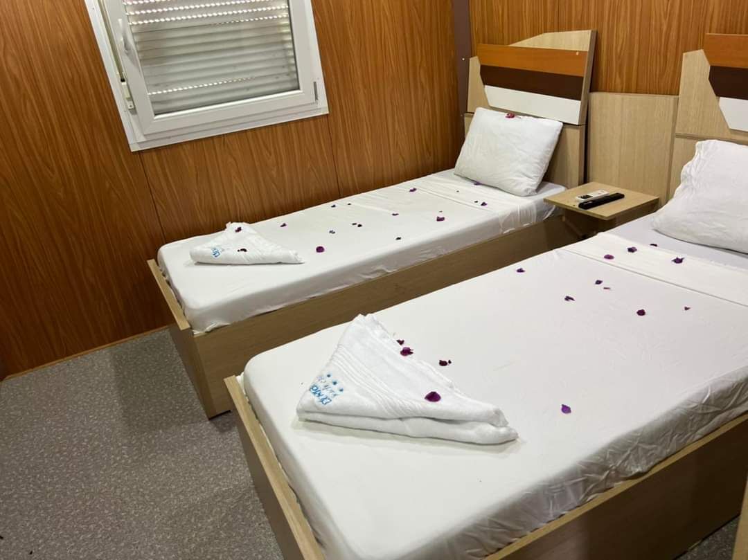 Dos camas individuales con sábanas blancas y pétalos de rosa, cabeceros de madera, una mesita de noche con teléfono y una ventana con persianas.