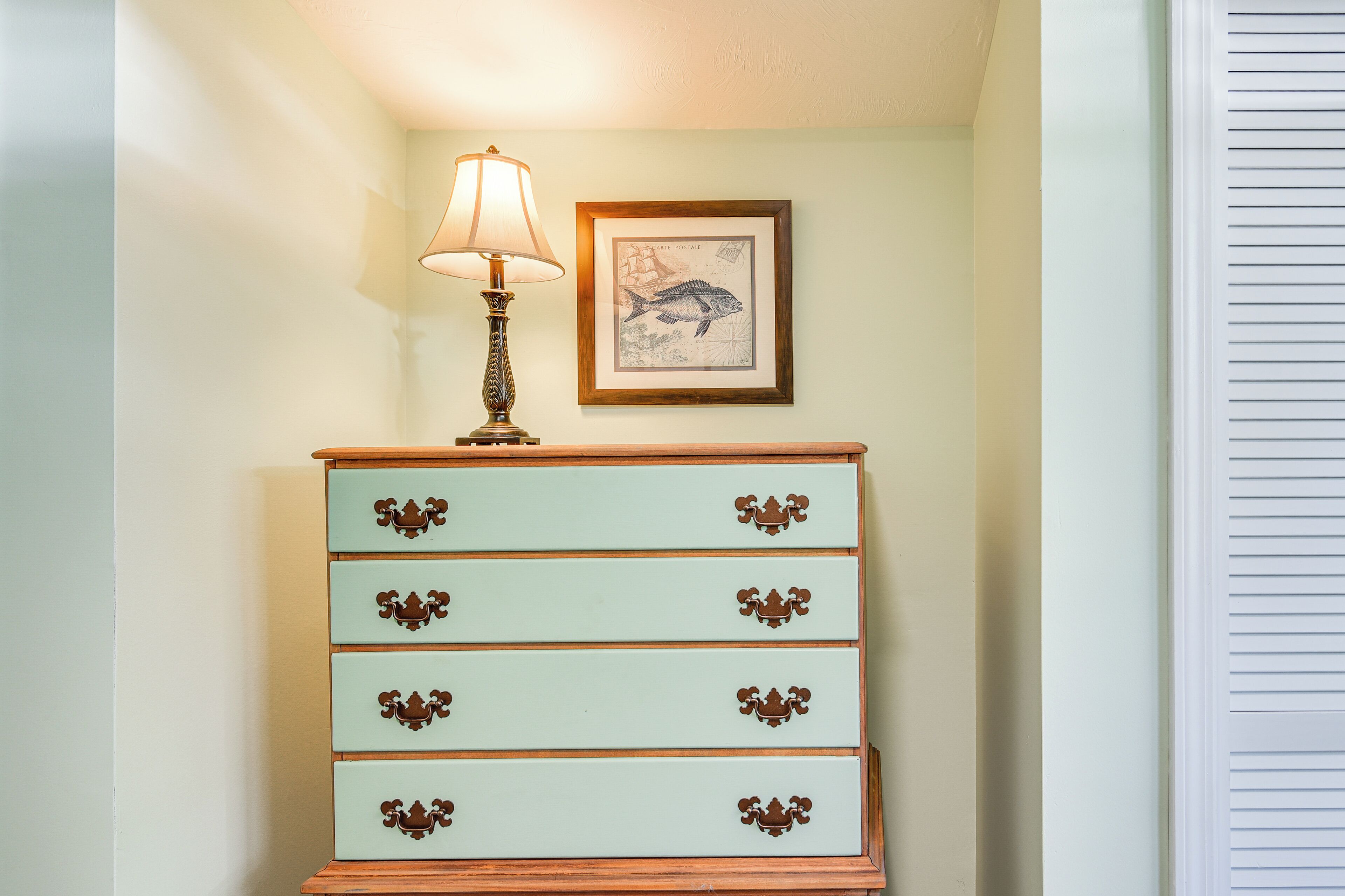 A light green dresser with ornate handles, a lamp, and a framed nautical illustration on the wall.
