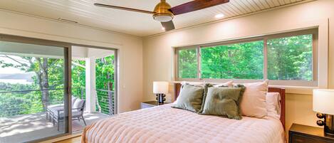 Ein Schlafzimmer mit einem großen Bett, einem Deckenventilator, einem Nachttisch, einer Lampe und Blick auf Grün durch das Fenster.