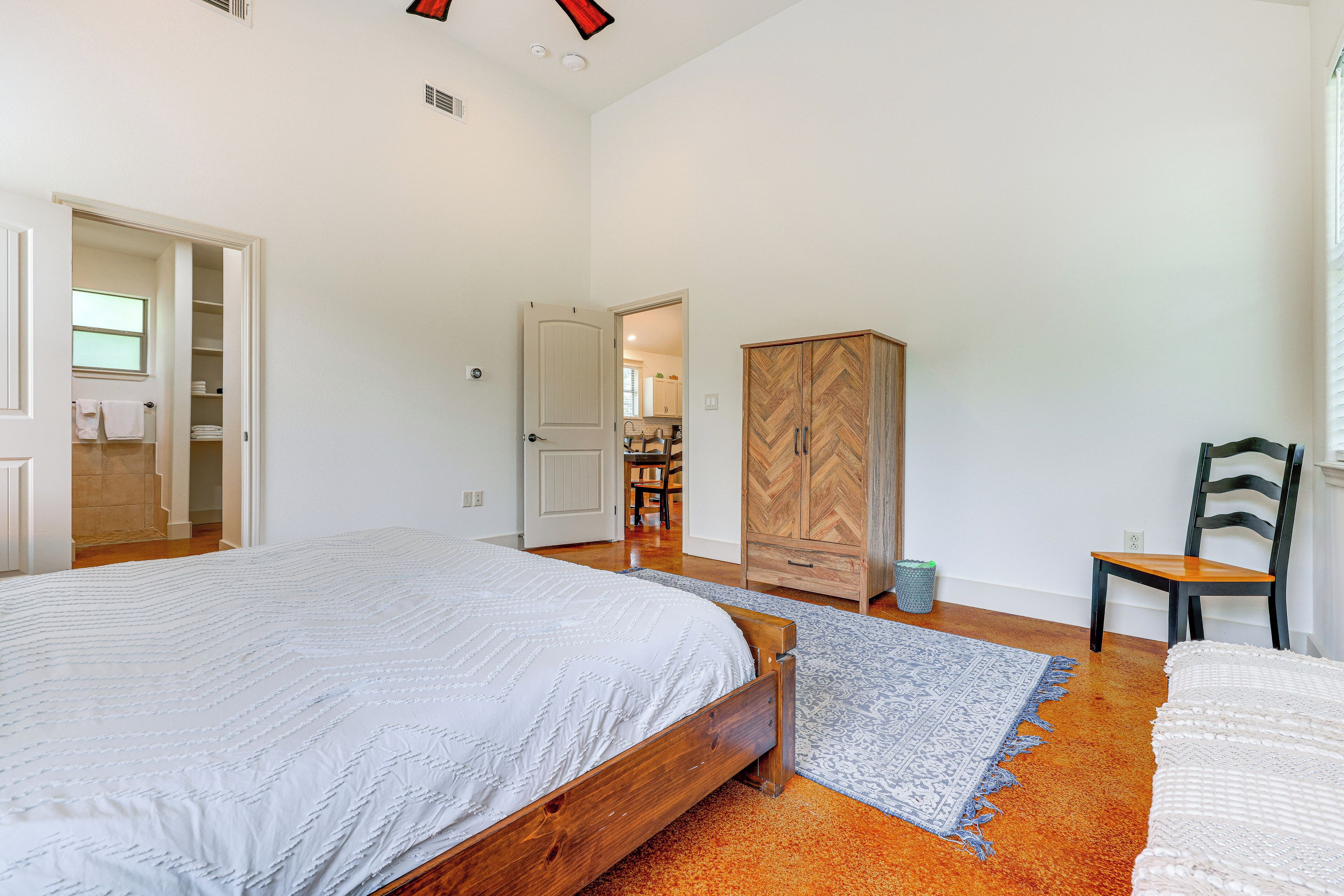 Un dormitorio con una cama grande, un armario y una silla. Hay una puerta que da a un baño y una cocina.