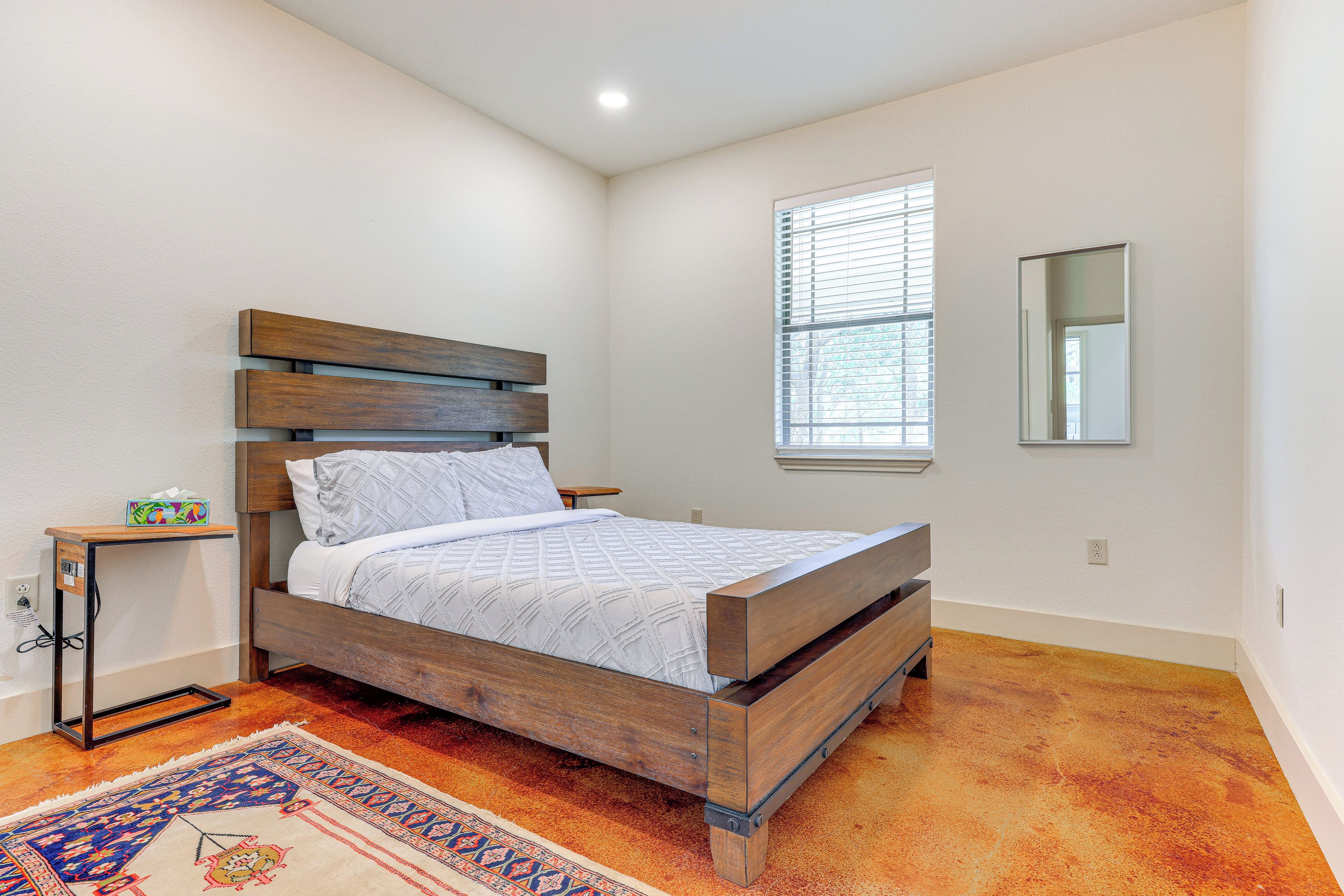 A bedroom with a wooden bed, a nightstand, a bedside table, a window with blinds, and a mirror on the wall.