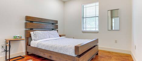 A bedroom with a wooden bed, a nightstand, a bedside table, a window with blinds, and a mirror on the wall.