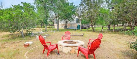 A circular stone fire pit with four red chairs around it, set in a grassy outdoor area with trees and a building in the background.