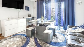 Una sala de estar moderna con comedor, televisor de pantalla plana en la pared y vistas de la ciudad a través de amplias ventanas con cortinas azules.