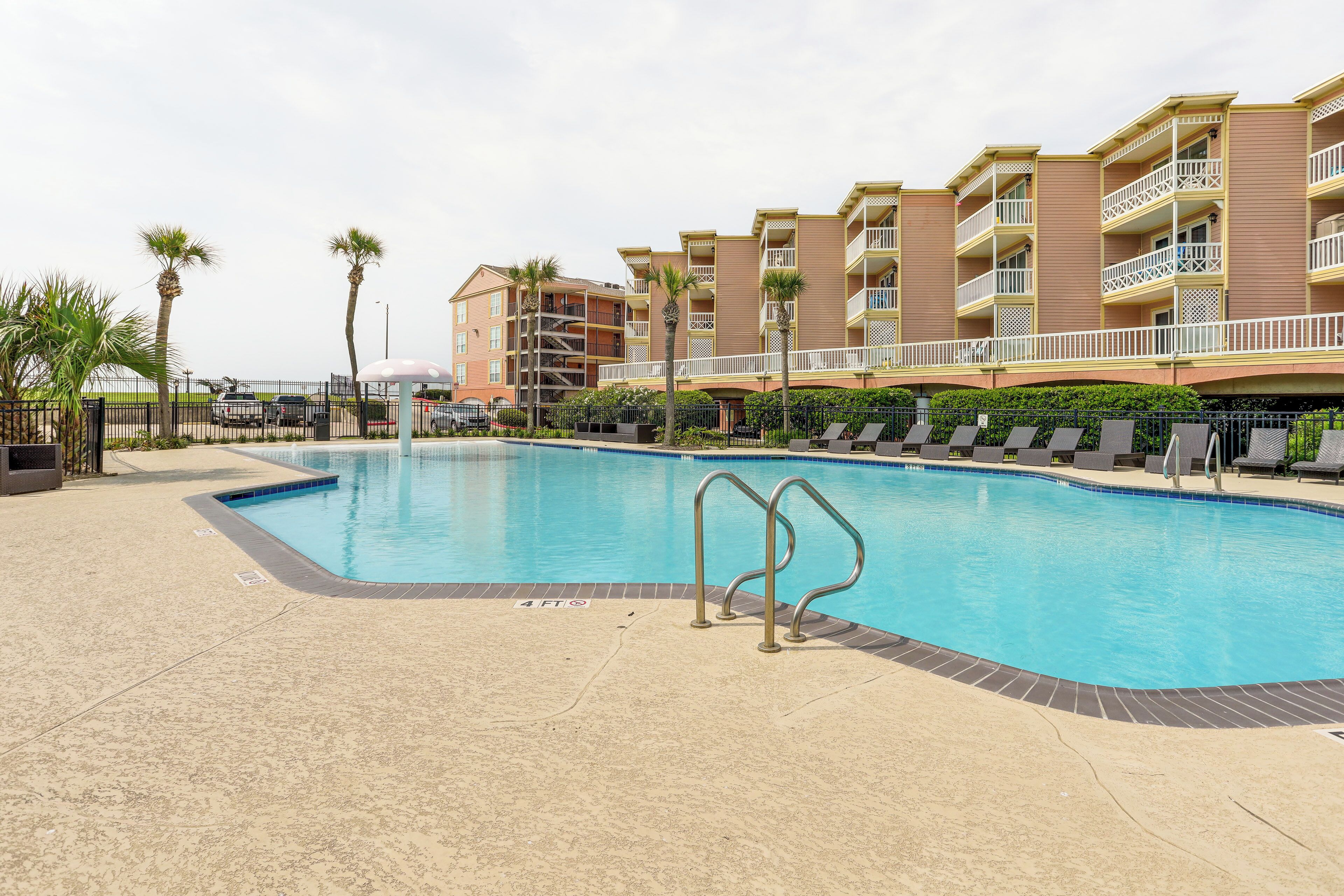 A swimming pool with a 4ft depth marker, surrounded by lounge chairs and palm trees, with a multi-story building in the background.