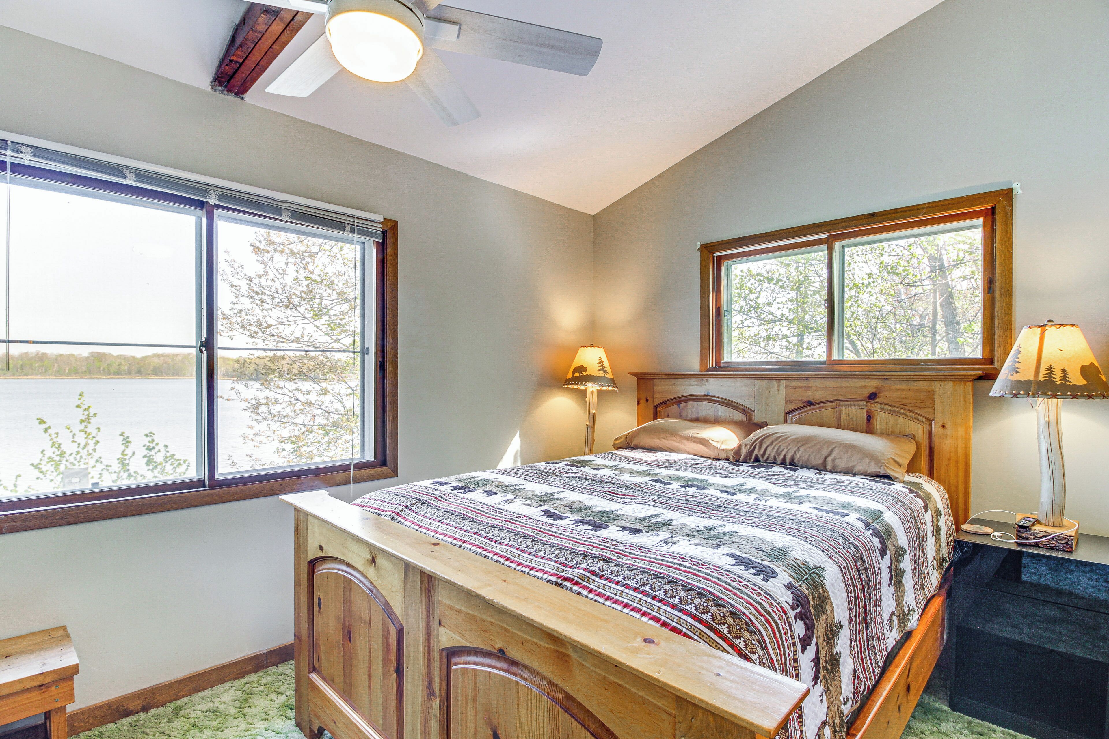 A bedroom with a large bed, two windows, a ceiling fan, and a nightstand with lamps.