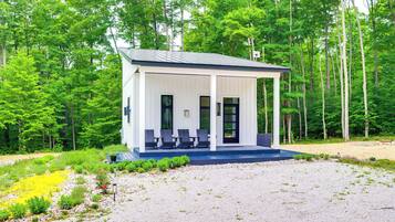 A small, single-story cabin with a covered porch and a few chairs, surrounded by a forest.