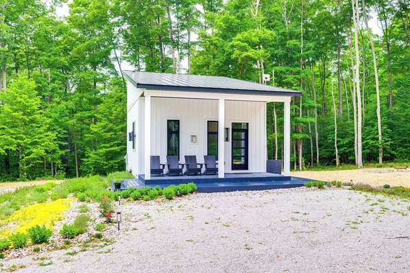 A small, single-story cabin with a covered porch and a few chairs, surrounded by a forest.