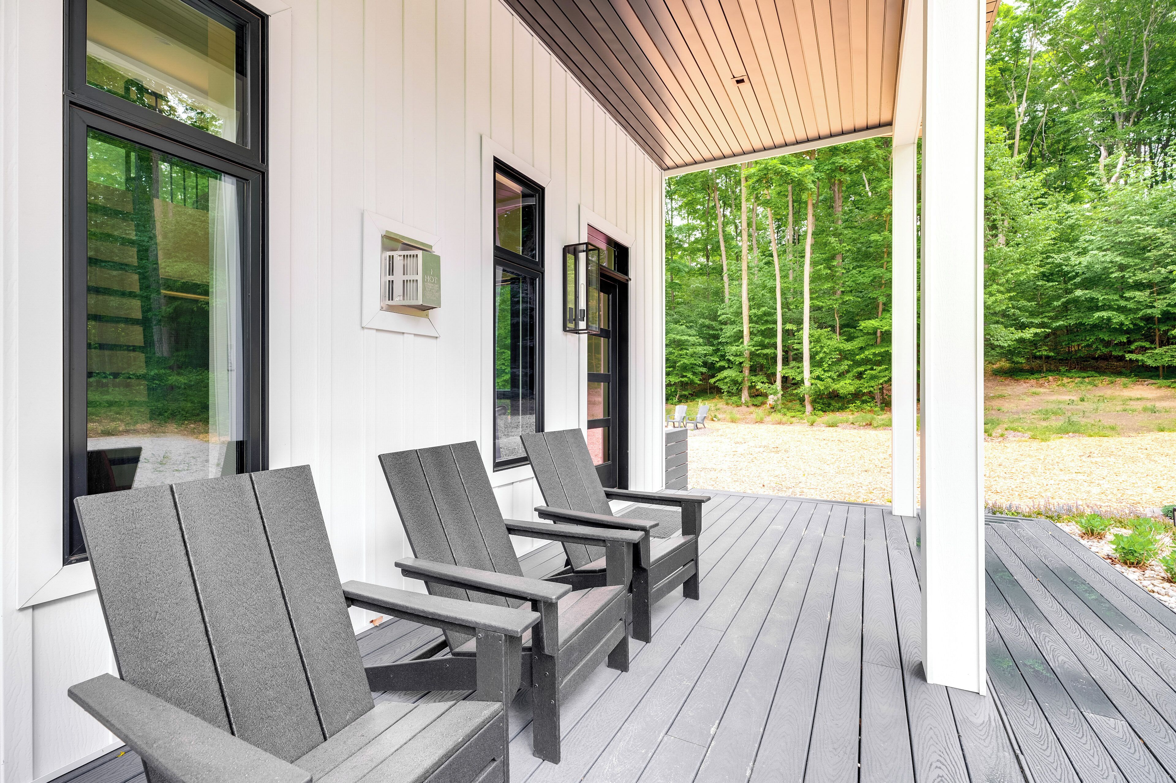 A covered outdoor seating area with Adirondack chairs, a wooden deck, and a view of a forested area.