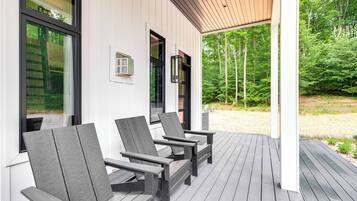 A covered outdoor seating area with Adirondack chairs, a wooden deck, and a view of a forested area.