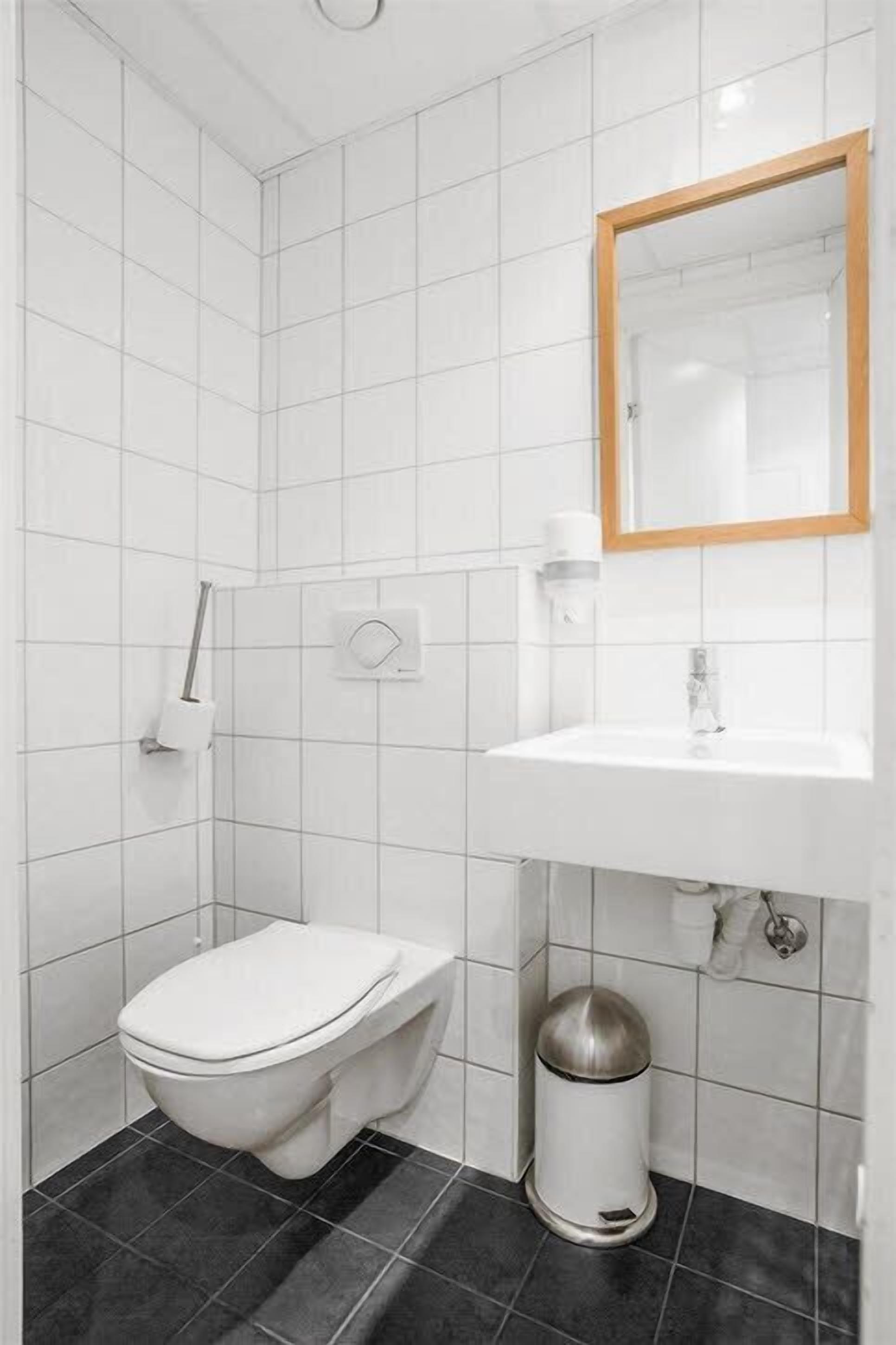 A small, modern bathroom with white tiles, a white sink, a mirror, and a toilet.
