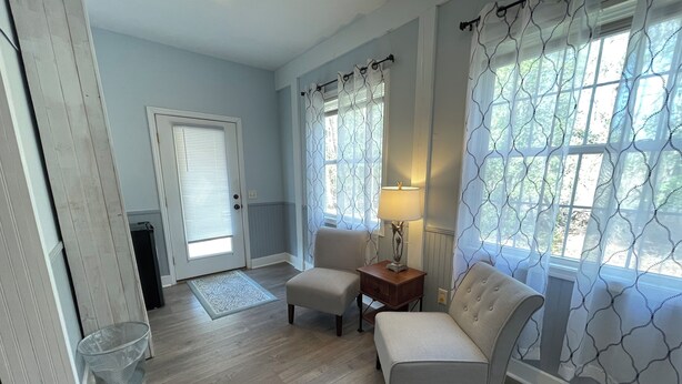 Une pièce avec un mur bleu clair, un plancher en bois, une porte blanche, une petite table avec une lampe, deux fauteuils et une fenêtre ornée de rideaux à motifs.