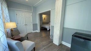 Une chambre d’hôtel de taille réduite comprenant un lit, un petit coin salon avec une chaise et une table, un réfrigérateur et une porte donnant sur une autre pièce.