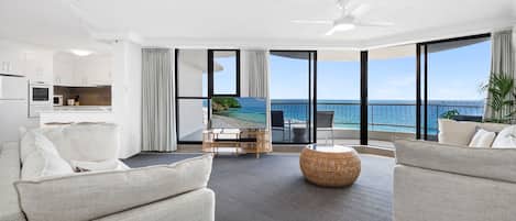 A modern living room with a view of the ocean, a balcony with outdoor seating, and a kitchen area.
