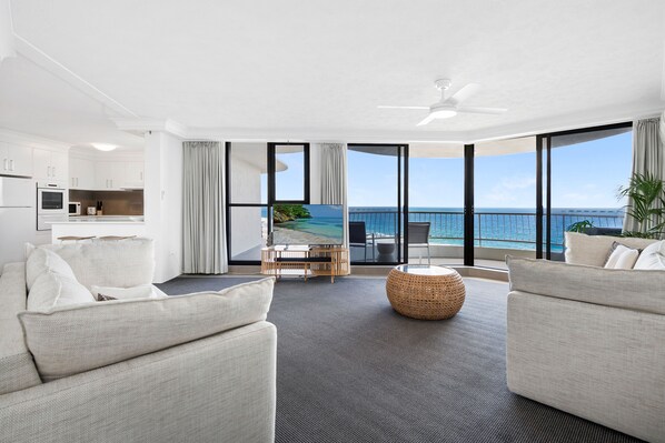 A modern living room with a view of the ocean, a balcony with outdoor seating, and a kitchen area.