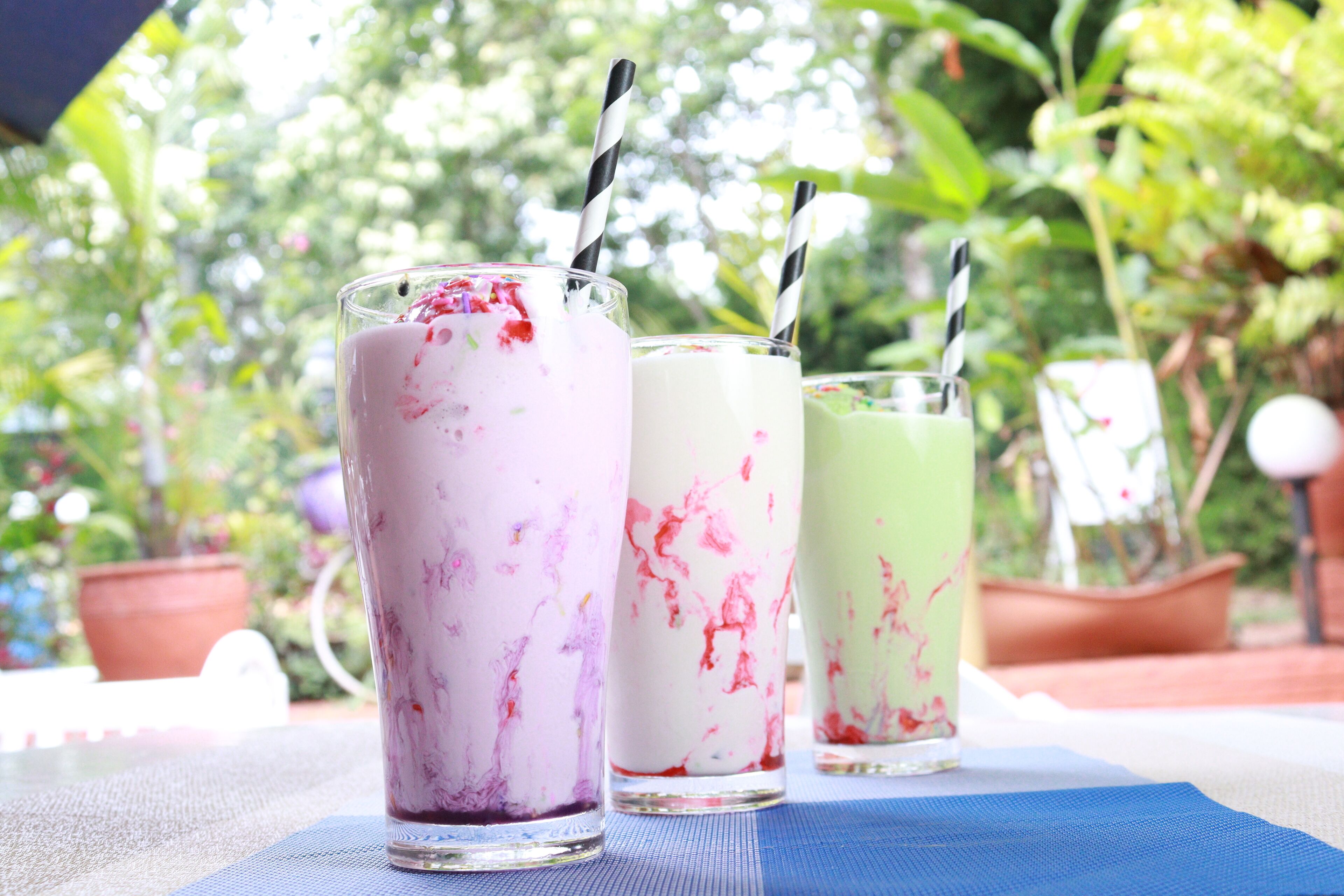 Tres vasos de bebidas coloridas con pajitas sobre una mesa cubierta con un mantel azul, al aire libre con follaje al fondo.