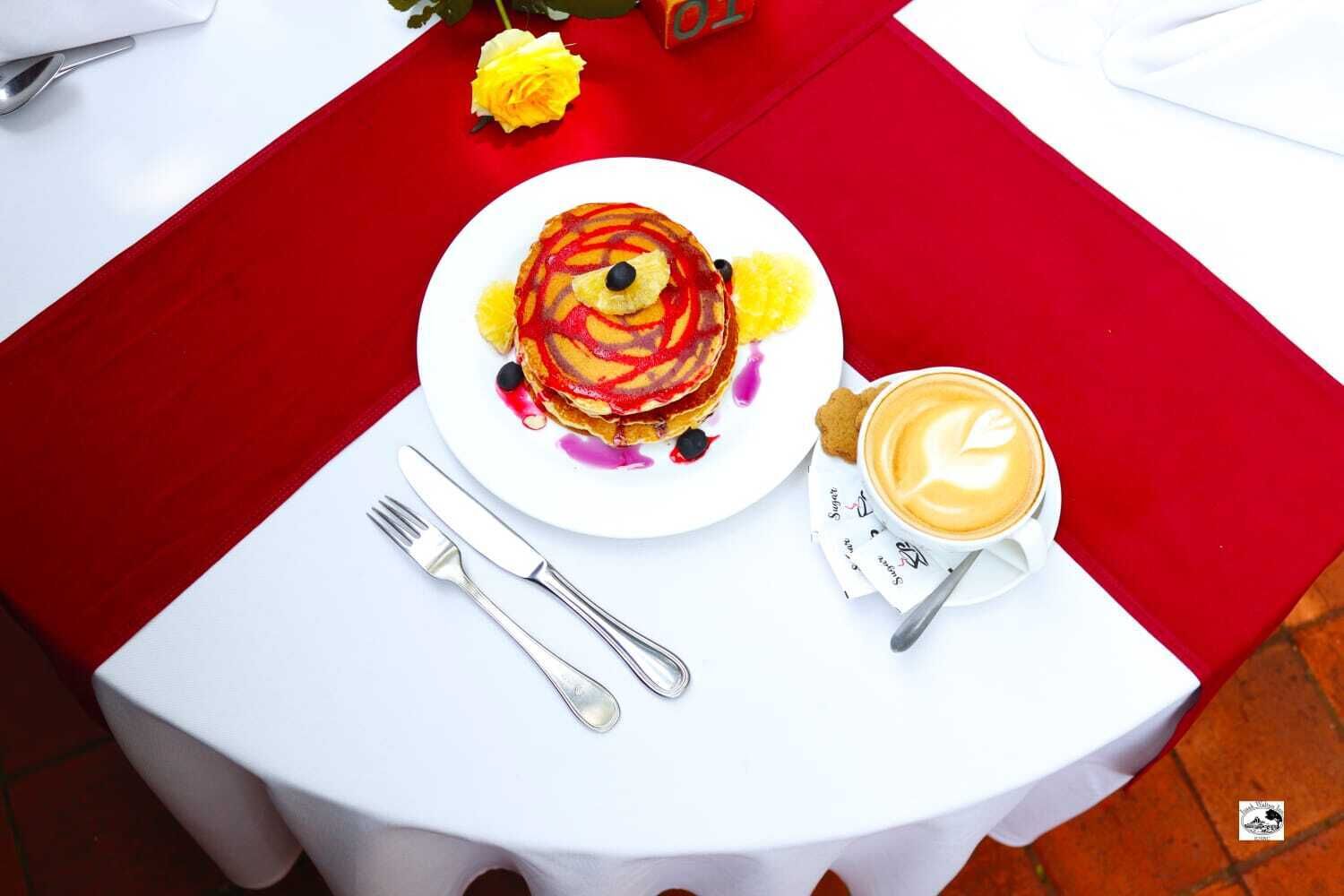 A breakfast setting with pancakes, coffee, and utensils on a white table with a red tablecloth.