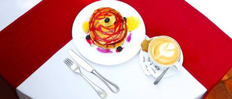 A breakfast setting with pancakes, coffee, and utensils on a white table with a red tablecloth.