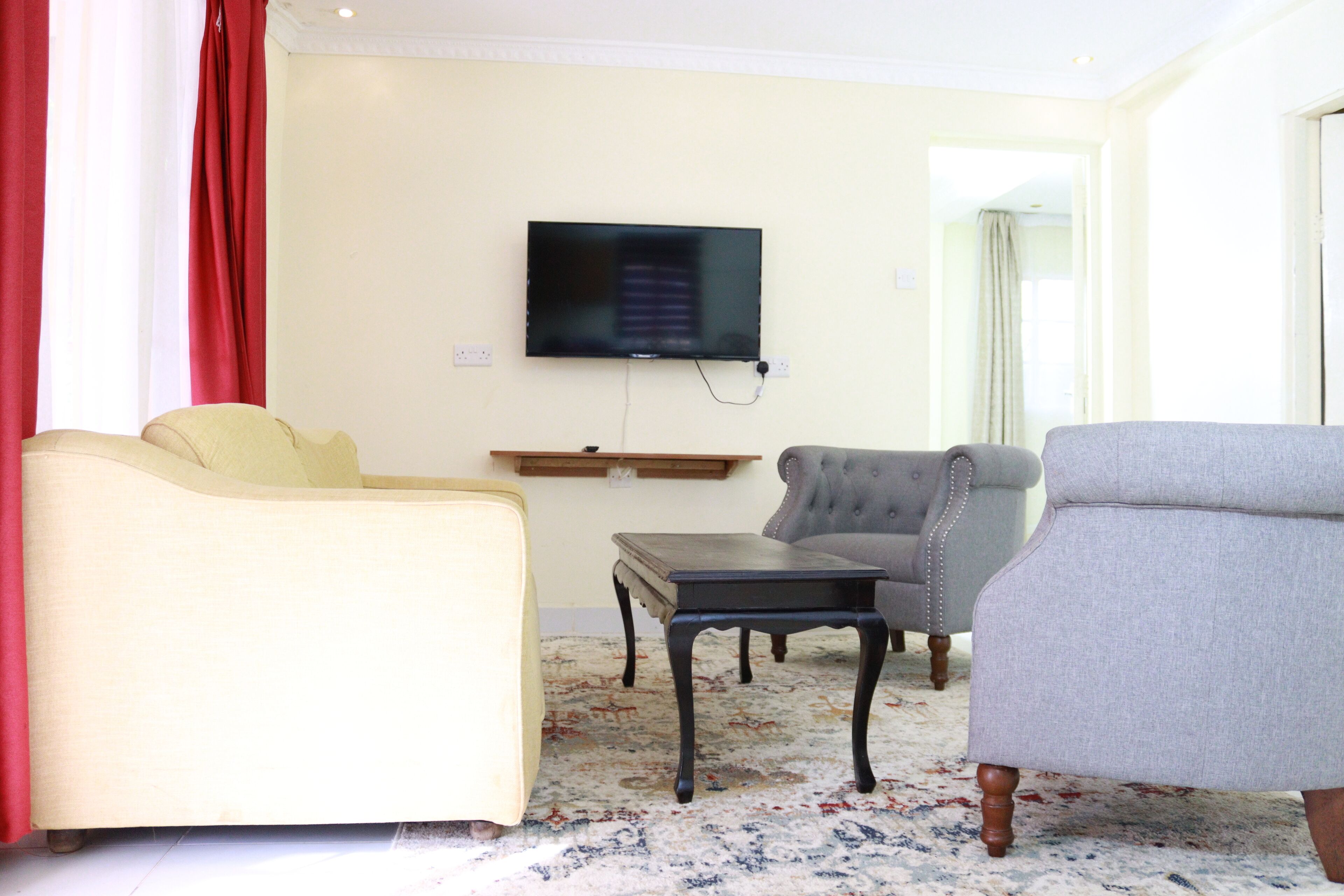 A living room with a flat-screen TV mounted on the wall, a beige sofa, a grey armchair, a small wooden table, and a patterned carpet.