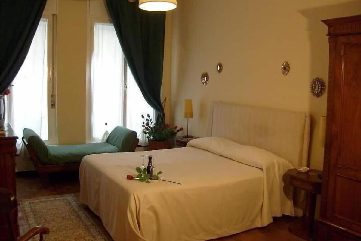 A bedroom with a bed, a chair, a nightstand, a lamp, and a window with curtains.