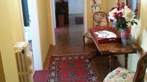 A hallway with a patterned rug, a wooden table with a chair, a clock, and a floral painting on the wall.