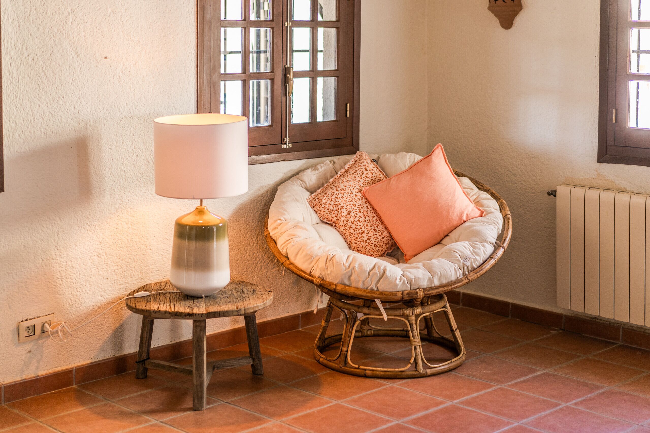 Une pièce chaleureuse avec un fauteuil en osier, une petite table avec une lampe et une fenêtre donnant sur un beau paysage.