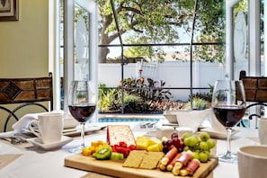 Une table de petit-déjeuner garnie d’une variété de fruits, de fromages et de biscuits, accompagnée de verres à vin, le tout disposé devant une grande fenêtre donnant sur un jardin.