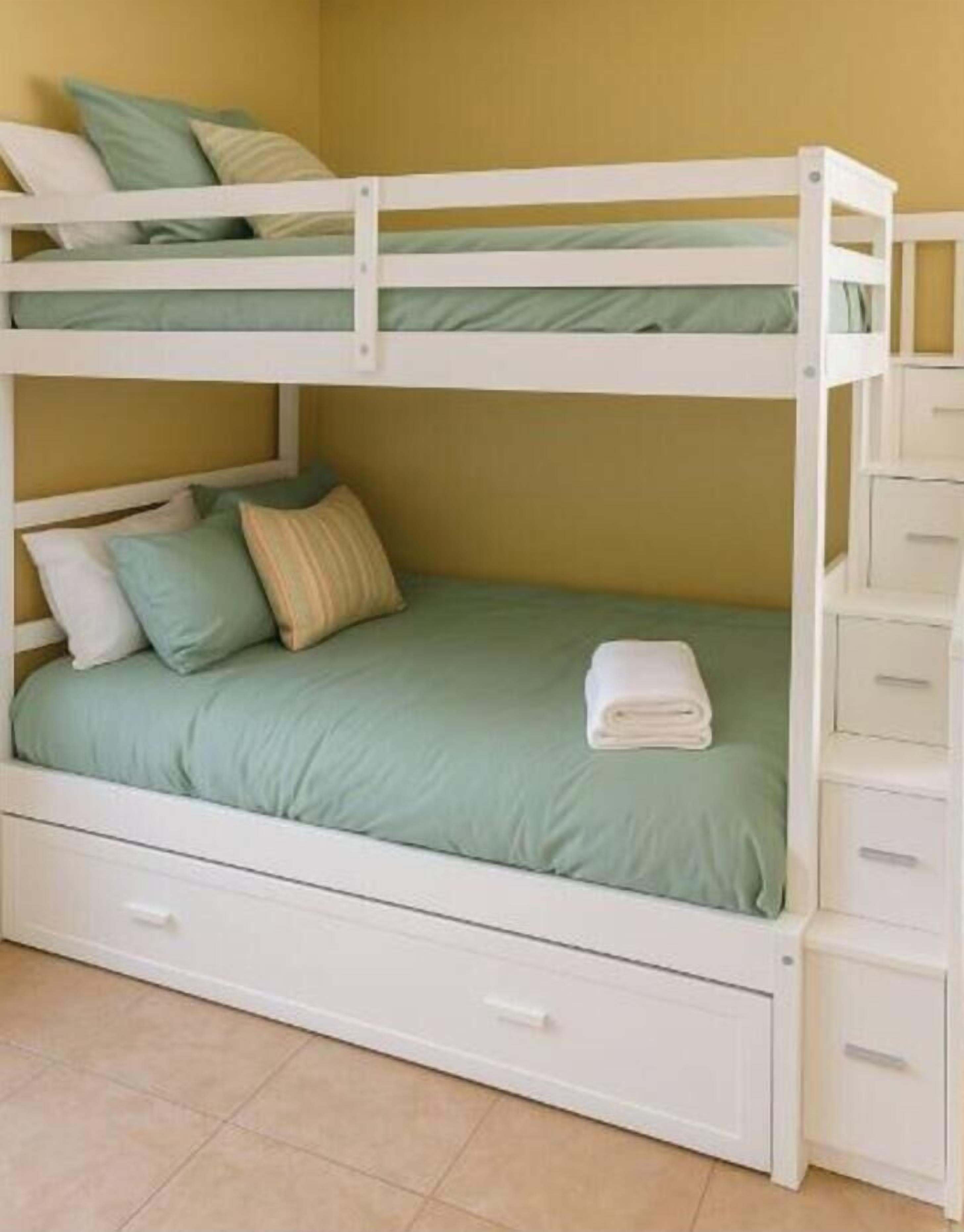 A bunk bed with a green mattress, white bedding, and a white drawer unit.