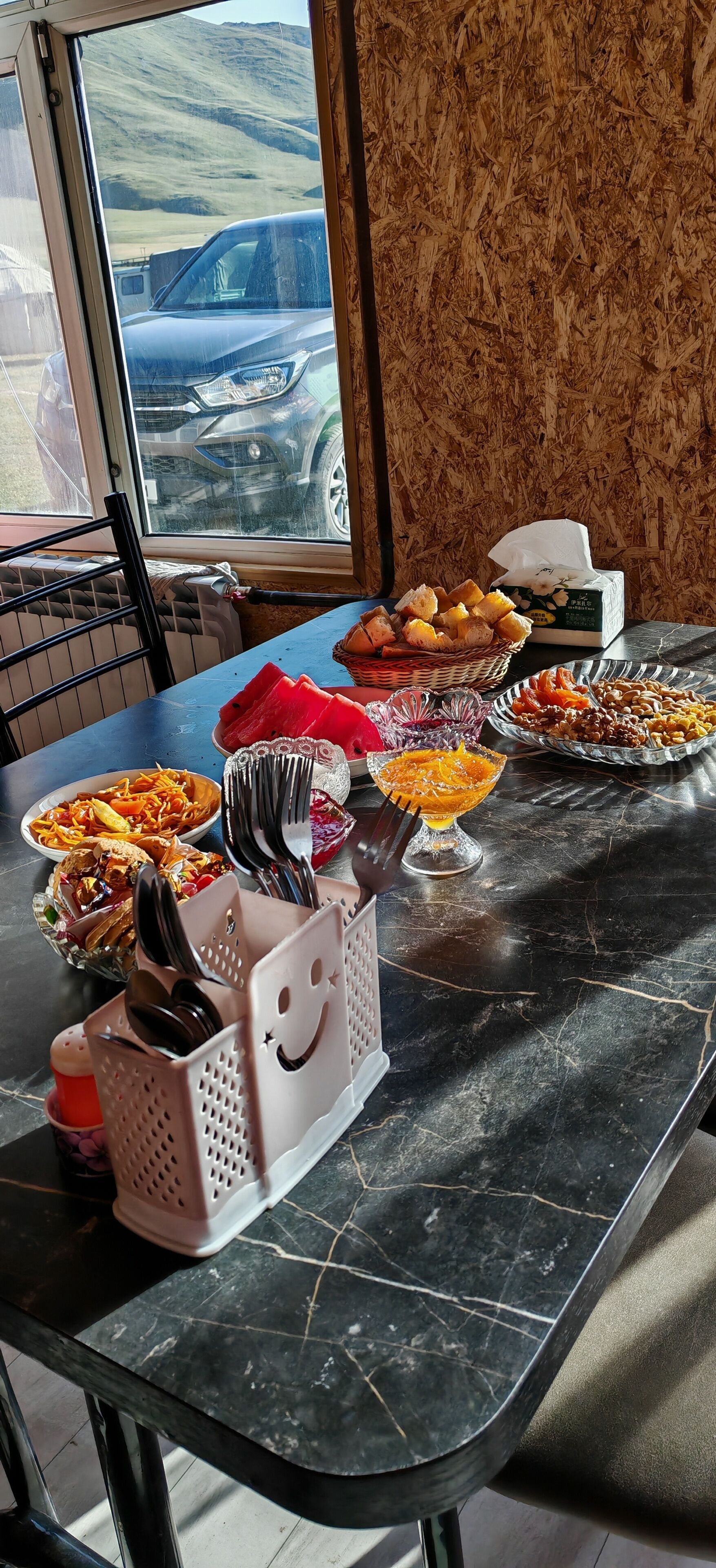 Uma mesa de jantar com diversos alimentos, utensílios, um cesto de pão e uma caixa de lenços.