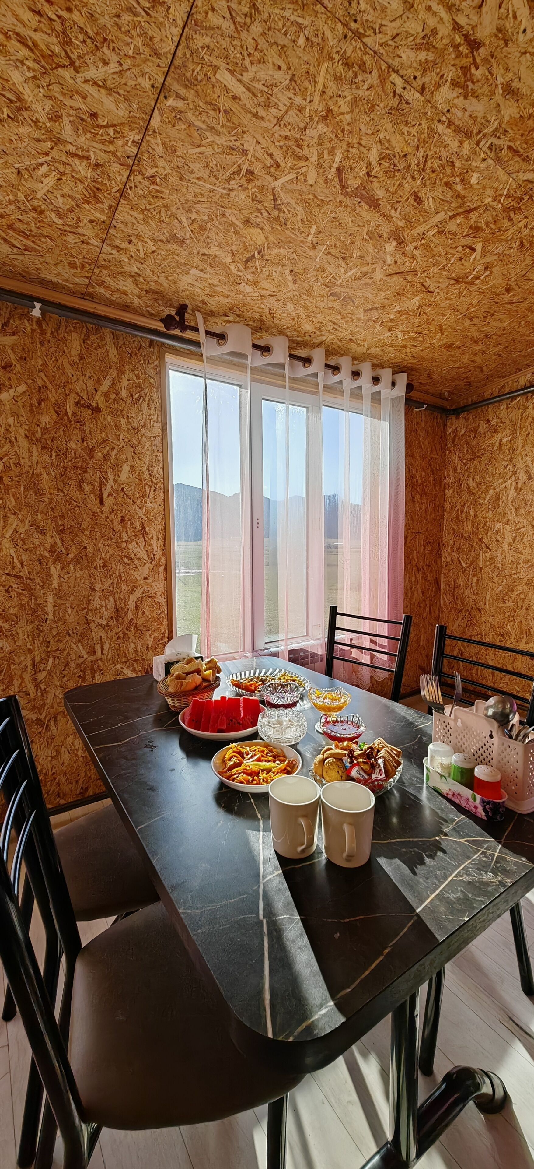 Ein Essbereich mit Tisch, gedeckten Sets, Stühlen, einem Fenster mit Vorhängen und Blick ins Freie.