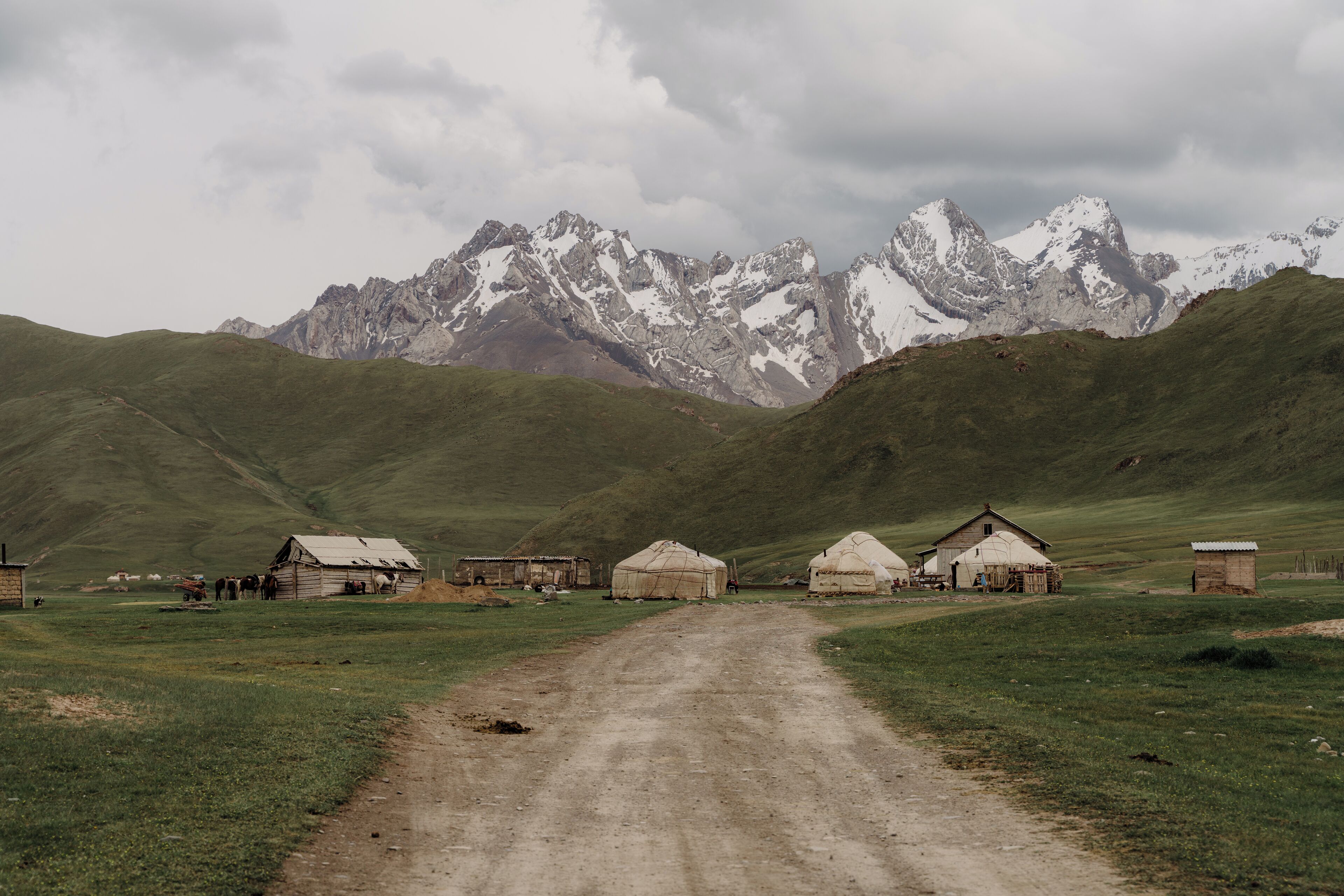 A rural landscape with a dirt road leading to yurts and a wooden structure, set against a backdrop of snow-capped mountains.