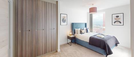 Un dormitorio con un armario grande, una cama con cabecero azul, una mesita de noche con lámpara y cuadros en las paredes.