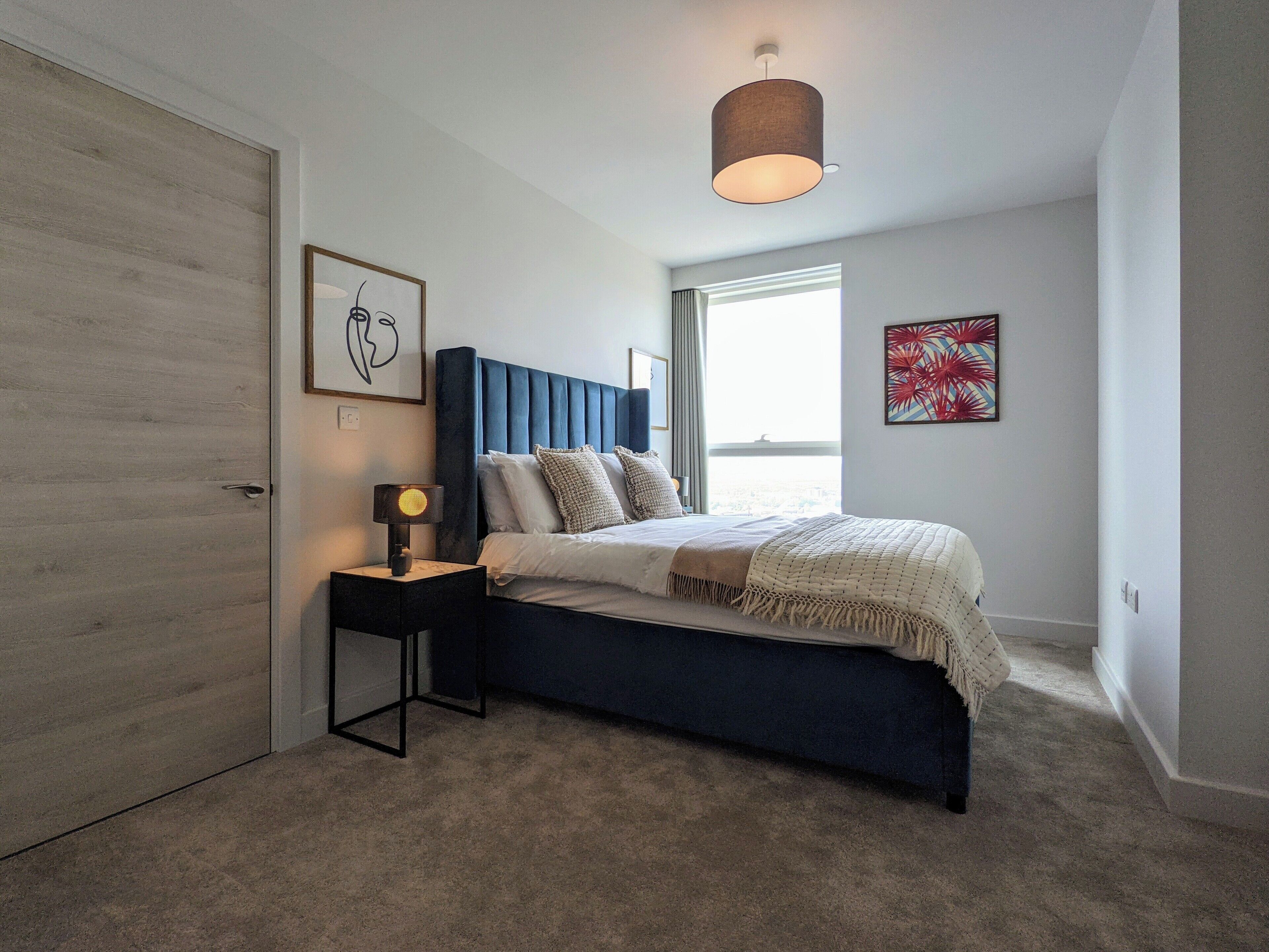 A bedroom with a bed, bedside table, lamp, artwork, and a window.