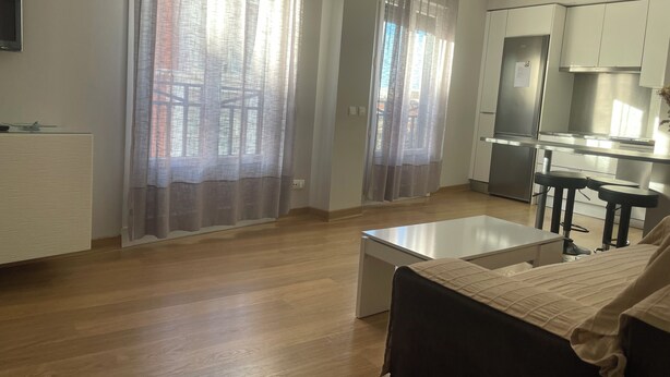A modern living room with a sofa, a coffee table, and a kitchen area with a refrigerator and bar stools. - Apartamento Centro muy Luminoso (Huelva)