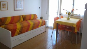 A small, bright room with a white sofa, a dining table with orange chairs, and a window with curtains. - Flat with big terrace in Grado (Grado Pineta)